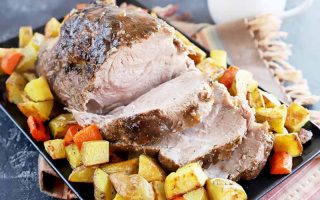 Horizontal image of a large piece of meat with slices on a serving platter surrounded by cubed potatoes and carrots.