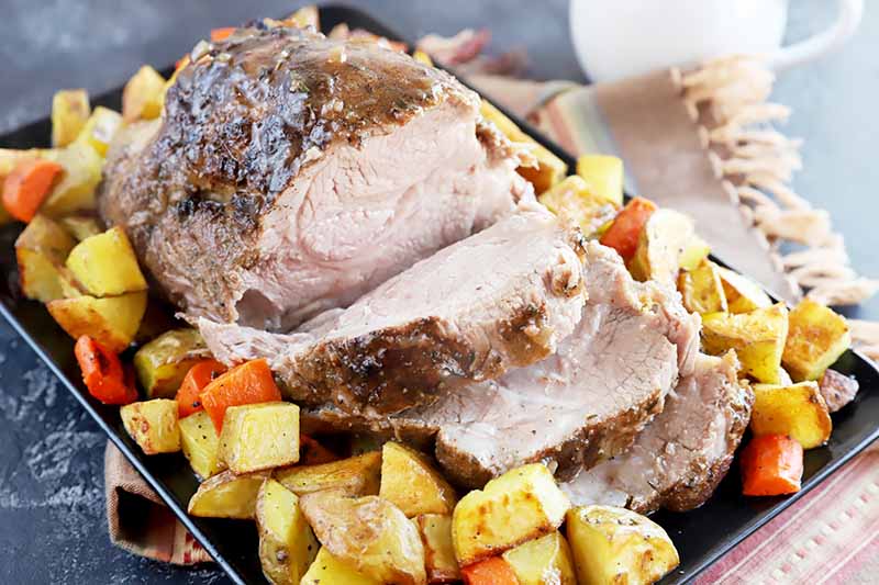 Horizontal image of a large piece of meat with slices on a serving platter surrounded by cubed potatoes and carrots.