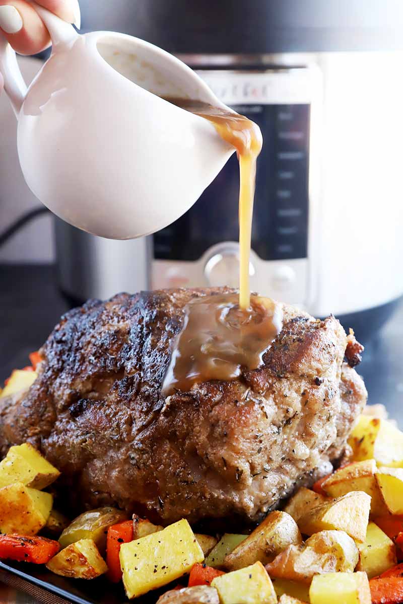 Vertical image of pouring gravy over a large piece of seared meat on a platter with chopped potatoes and carrots.