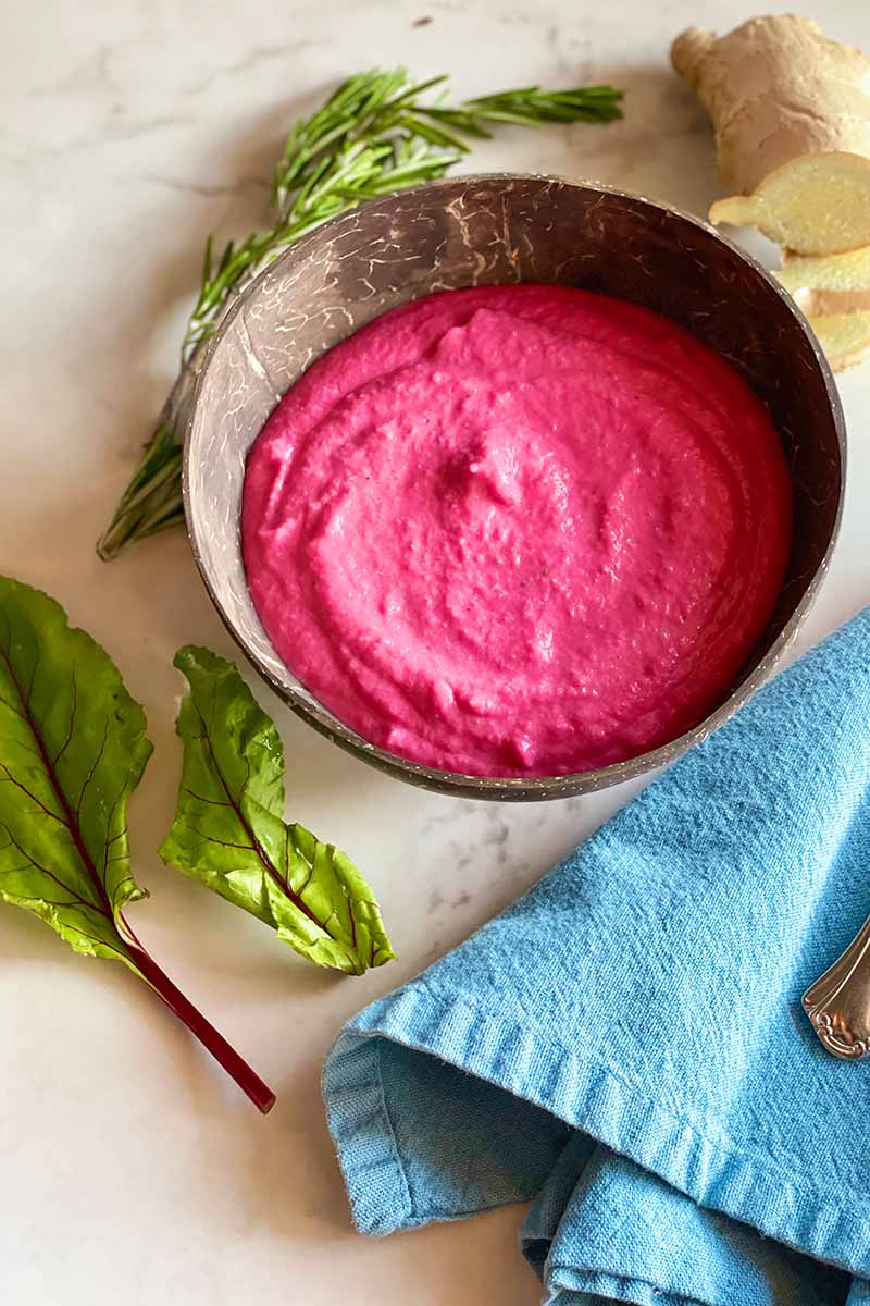 Vertical image of a wooden bowl filled with a bright pink puree next to herbs, ginger, and a blue towel.