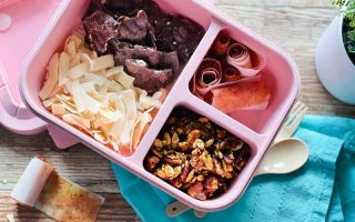 Horizontal image of a pink container with compartments filled with assorted food next to a blue towel.