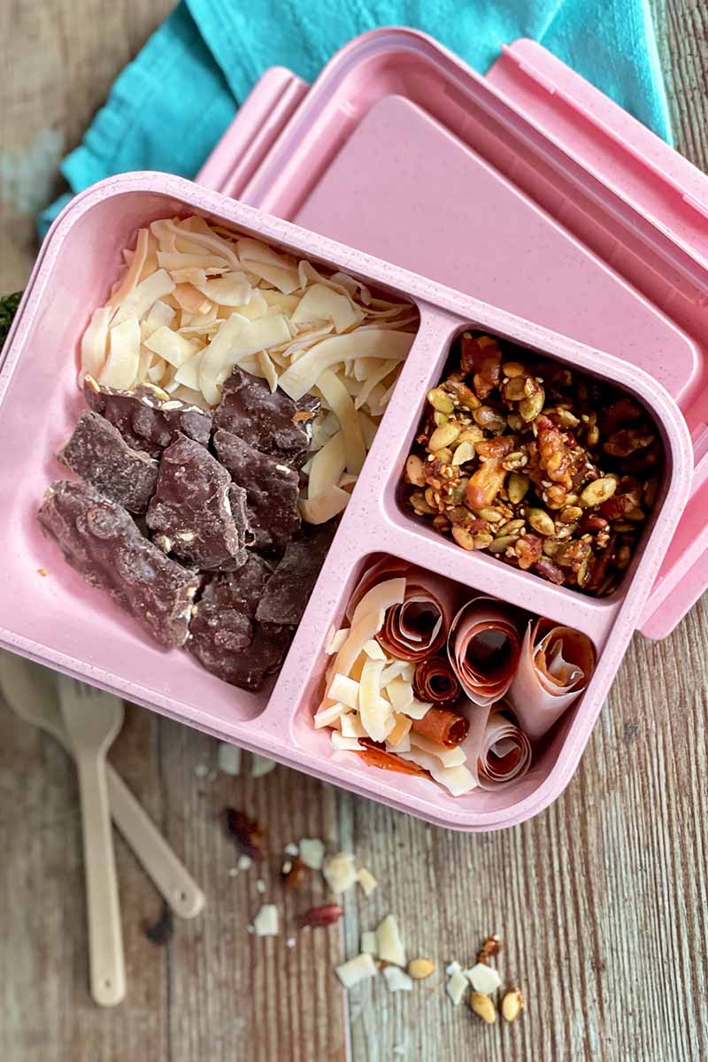 Vertical image of a pink container with compartments filled with assorted food next to a blue towel and plastic spoons.