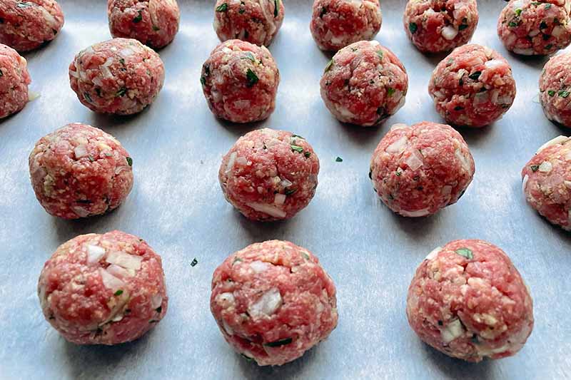 Horizontal image of round mounds of raw ground beef with seasonings on a baking sheet.