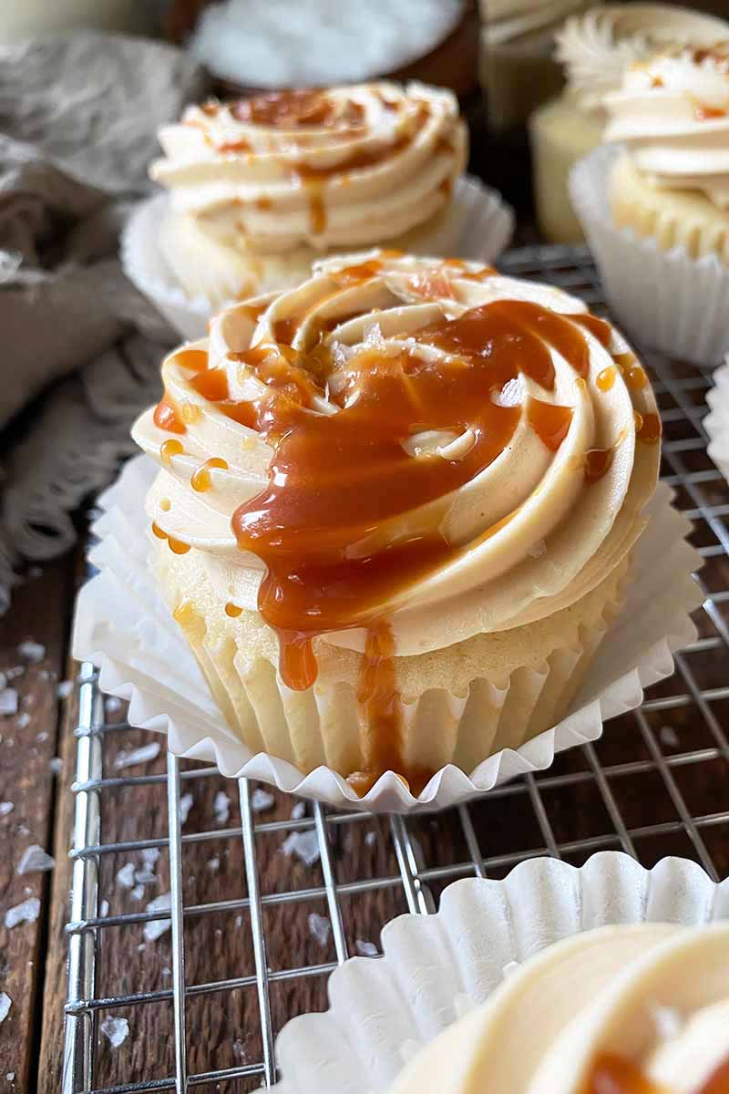 Vertical image of a thick brown sauce trailing down the side of a frosting cupcake on a cooling rack.