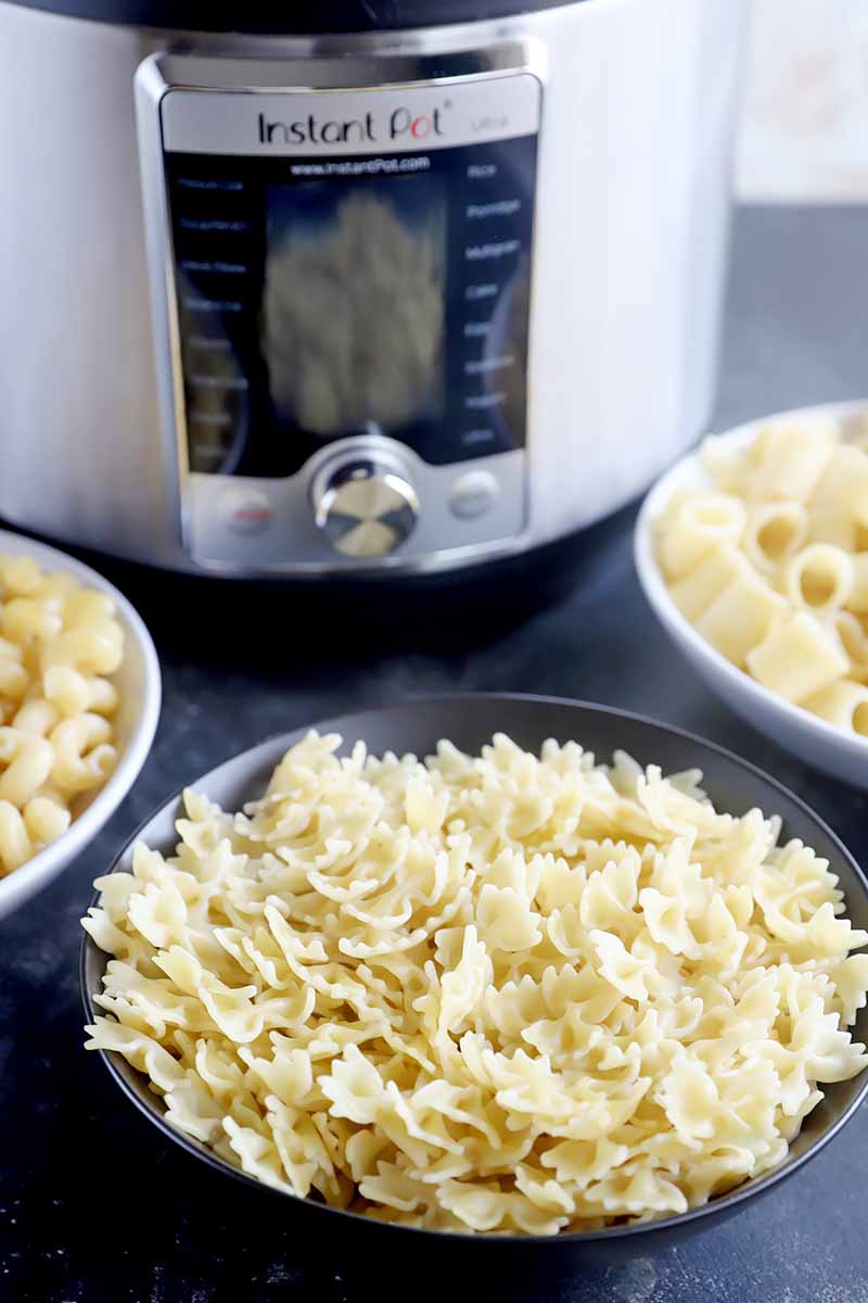 Vertical image of bowls of macaroni, rigatoni, and farfalle in front of a kitchen appliance.