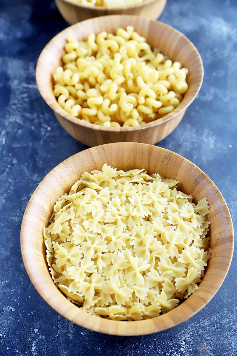 Vertical image of farfalle and macaroni in wooden bowls.