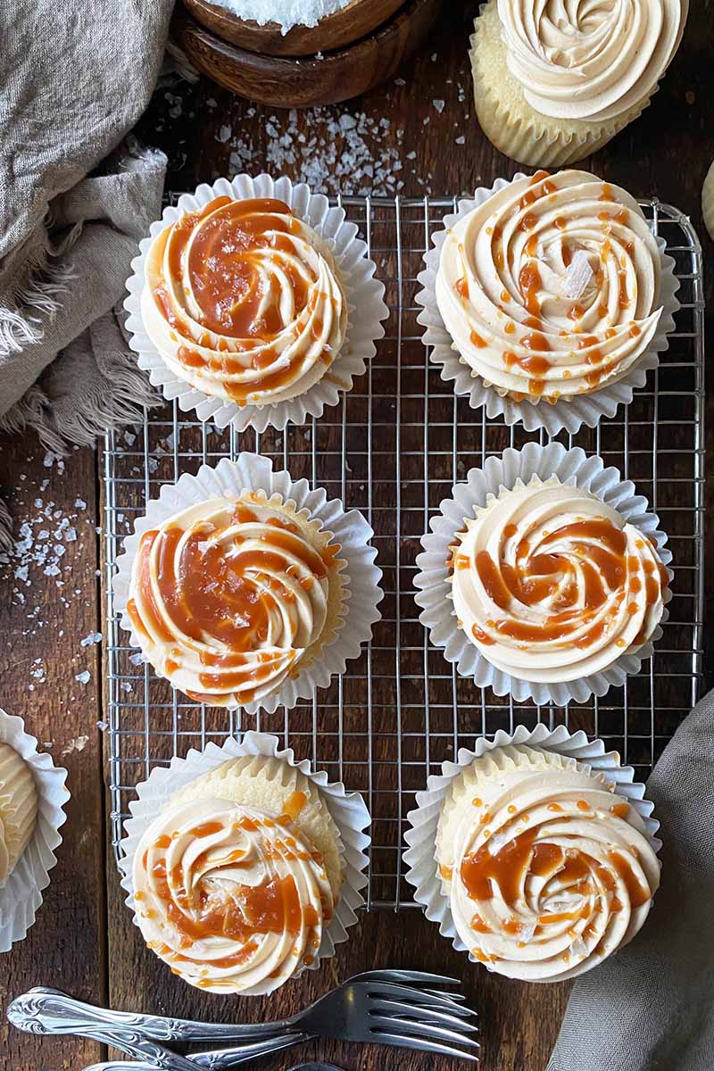 Vertical image of two rows of cupcakes with swirled buttercream drizzled with a brown sauce on a cooling rack next to scattered salt.