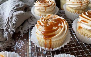 Horizontal image of rows of cupcakes with swirled buttercream and a drizzled brown sauce on a cooling rack next to a brown towel.