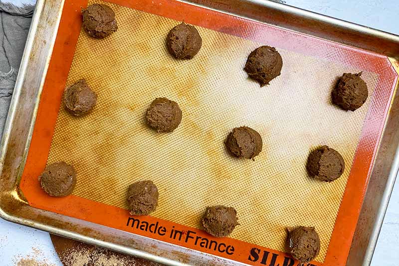 Horizontal image of portioned dark brown dough mounds on a baking sheet lined with a silicone mat.