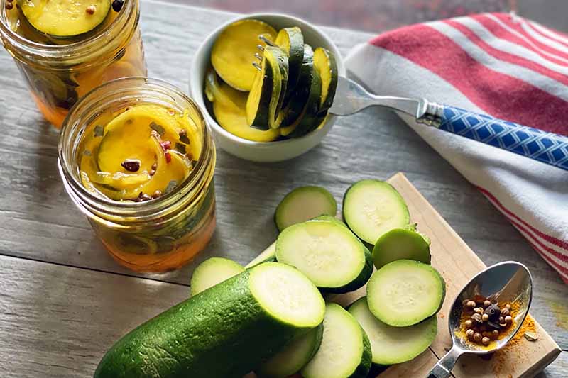 Horizontal image of slices of green vegetables on a wooden cutting board next to jars of pickles and a towel.
