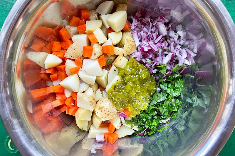 Horizontal image of mixed prepped vegetables, condiments, and herbs in a metal bowl.