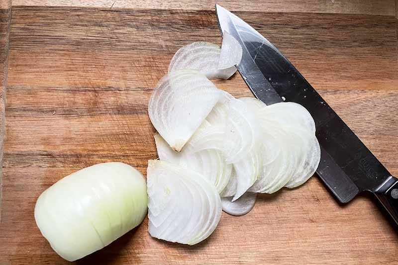 Horizontal image of sliced and halved onions next to a knife on a wooden cutting board.