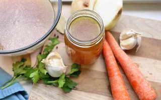Horizontal image of a jar and a bowl of light brown soup next to carrots, garlic, herbs, and onions on a wooden cutting board.