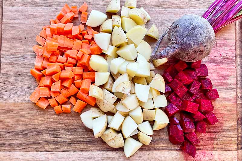 Horizontal image of cubed carrots, potatoes, and beets on a wooden cutting board.