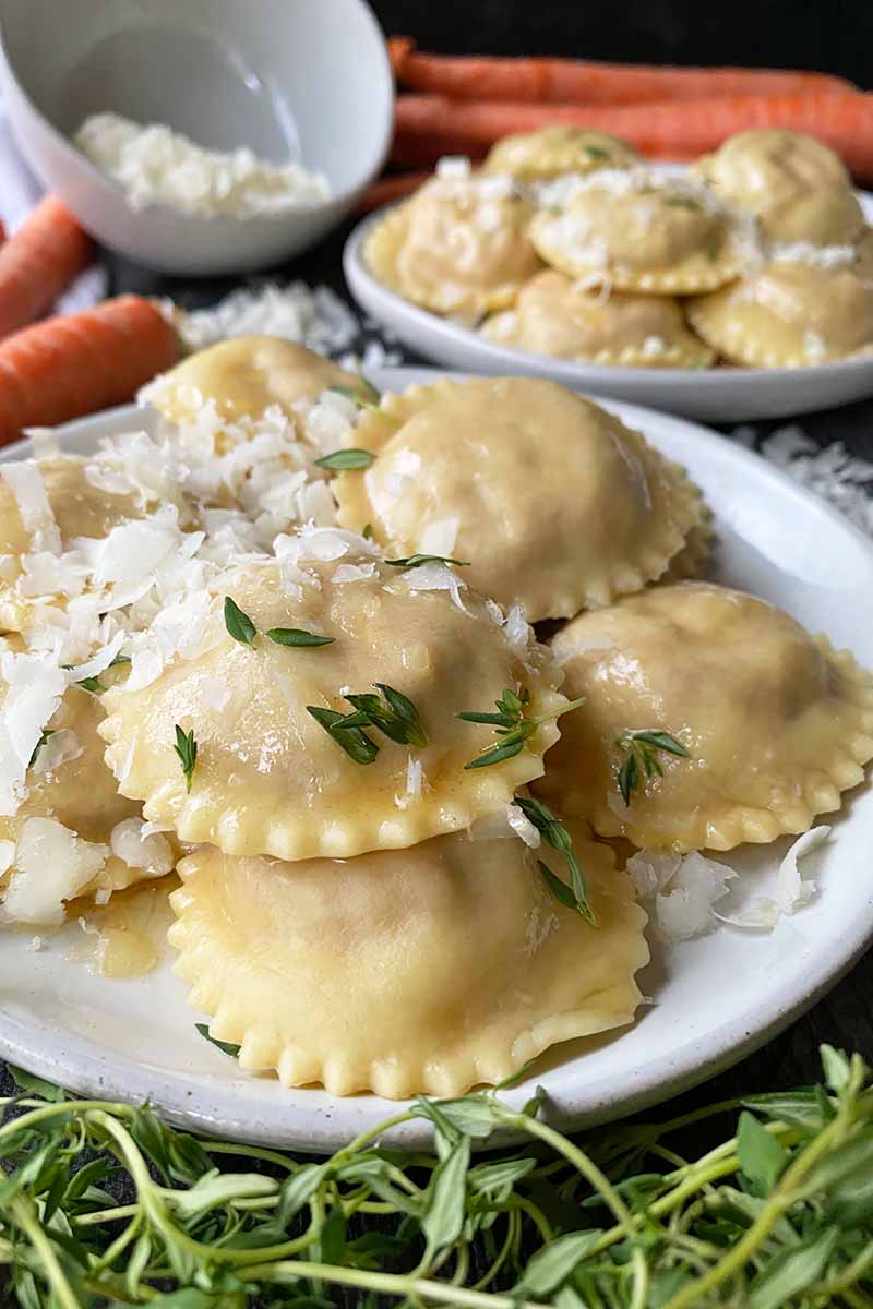 Vertical image of two plates with ravioli topped with cheese and herbs next to more grated cheese, herbs, and vegetables.