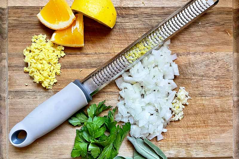 Horizontal image of a zester next to lemons, chopped onions, and fresh herbs.