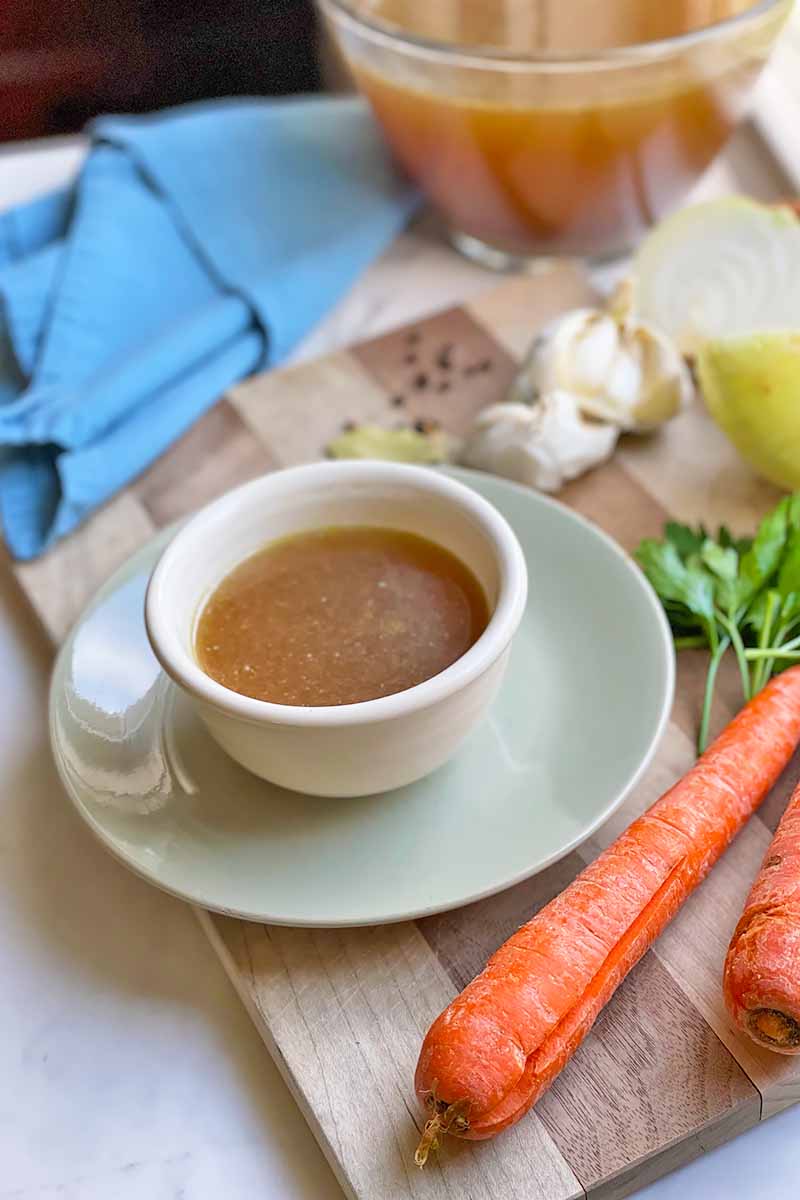 Vertical image of a bowl with light brown soup on a plate on a wooden cutting board next to a blue towel, carrots, garlic, and herbs.