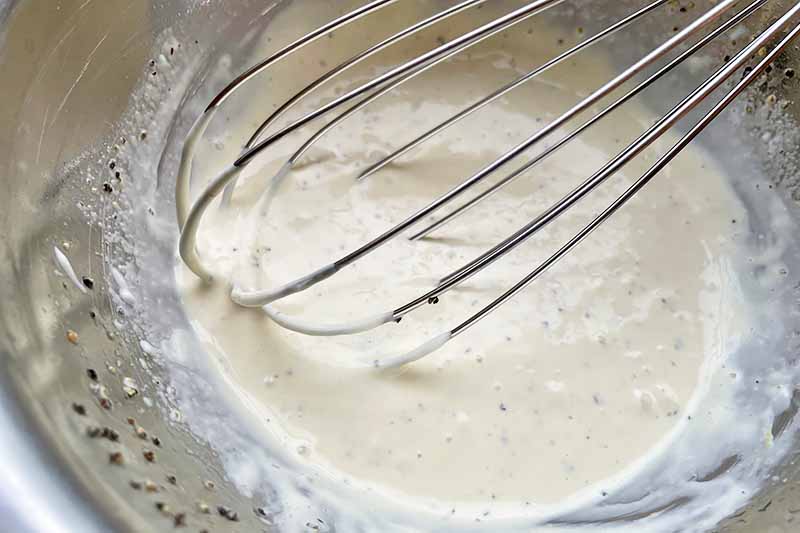 Horizontal image of a whisk mixing a creamy dressing together in a metal bowl.