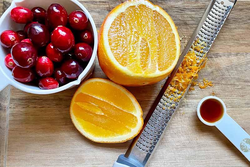 Horizontal image of a bowl of round red fruit, an orange, zest, a zester, and a teaspoon of vanilla.
