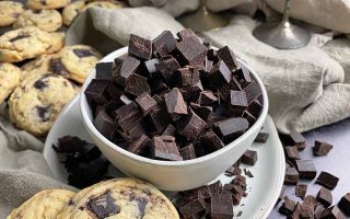 Horizontal image of a bowl filled with dark brown chunks of candy on a plate next to a tan towel and cookies.