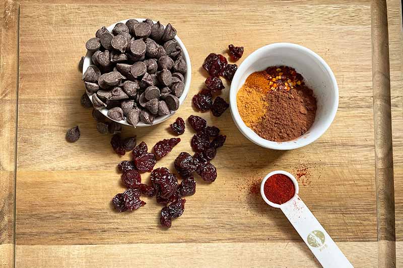 Horizontal image of a bowl of chocolate chips, a bowl of spices, scattered dried fruit, and a teaspoon of sweetener on a wooden cutting board.