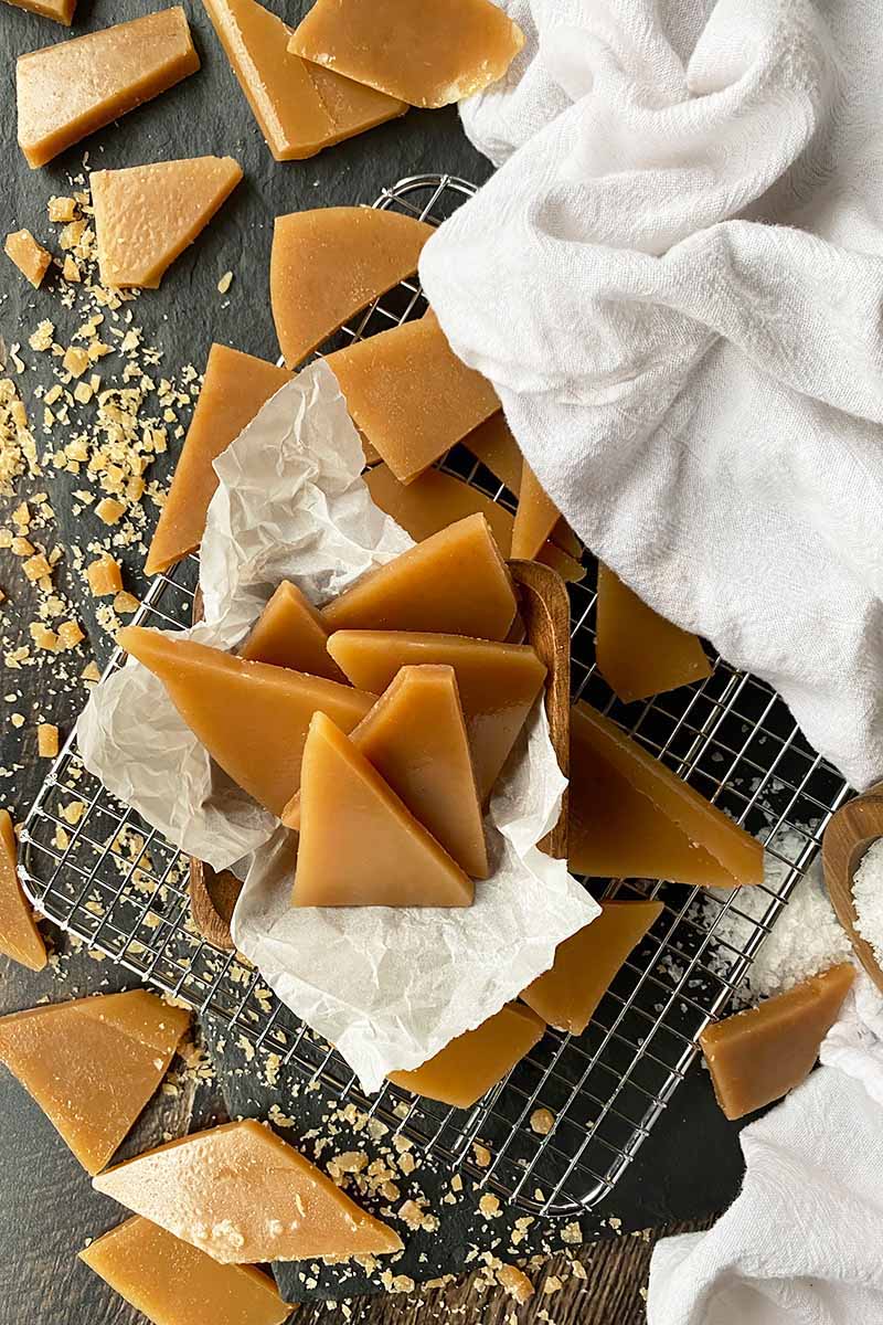 Vertical image of scattered toffee pieces on a dark surface with parchment paper, a white towel, and a cooling rack.