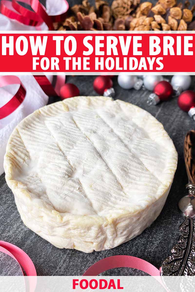 Vertical image of a whole wheel of cheese on a slate board next to ribbon, ornaments, and pine cones, with text on the top and bottom of the image.