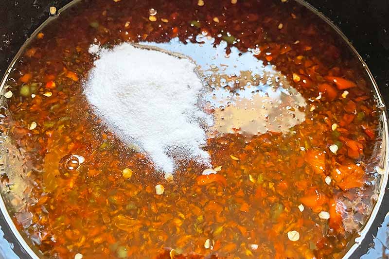 Horizontal image of powdered pectin on top of a chunky red and green liquid mixture in a pot.
