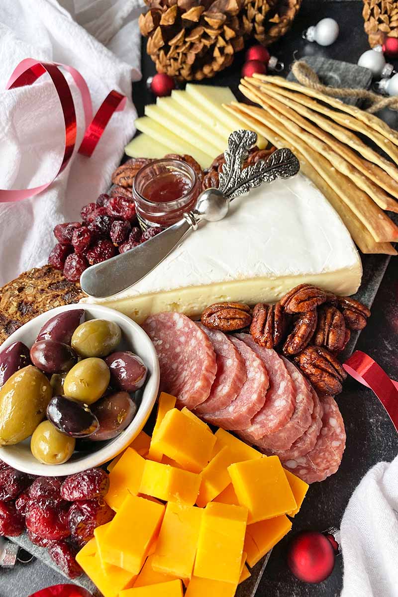 Vertical image of a wedge, slices, and cubes of cheese next to olives, salami, and crackers with jam and a knife.