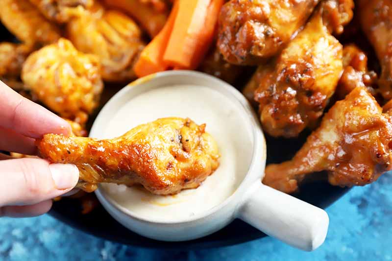 Horizontal image of dipping a drumette in a bowl of ranch dressing surrounded by wings and carrot sticks.