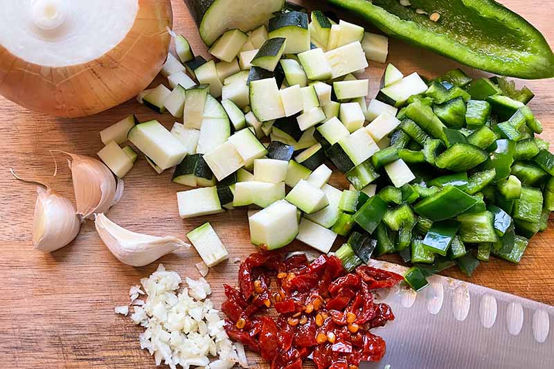 Horizontal image of assorted chopped vegetables and seasonings on a wooden cutting board next to a chef's knife.
