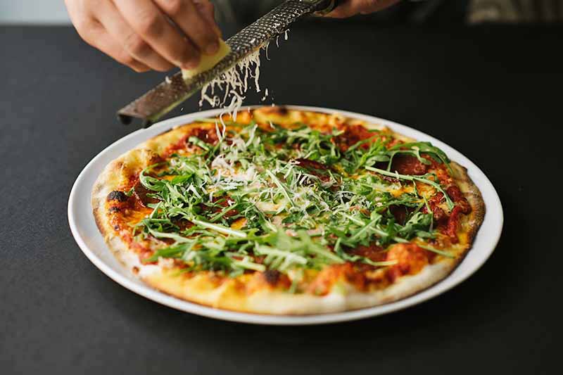 Horizontal image of grating cheese over a flatbread topped with arugula.