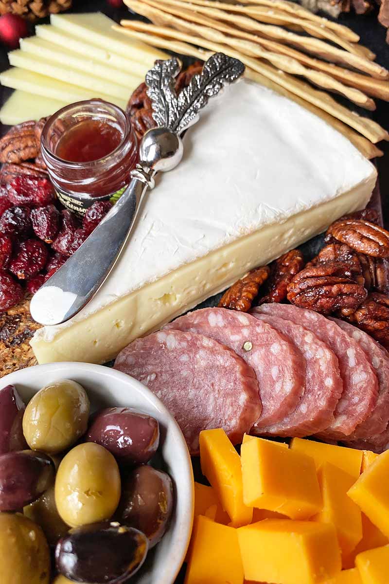 Vertical image of a wedge, slices, and cubes of cheese next to olives, salami, and crackers with jam and a knife.