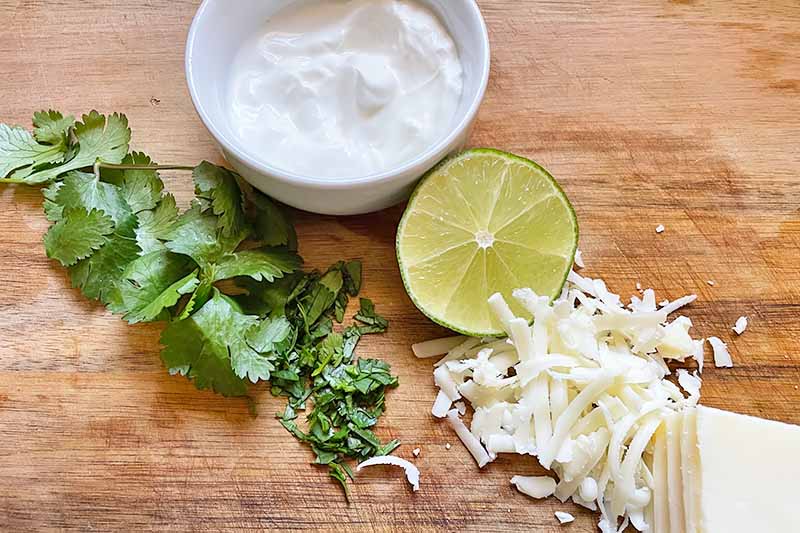 Horizontal image of chopped cilantro, sliced lime, shredded cheese, and a bowl of sour cream on a wooden surface.