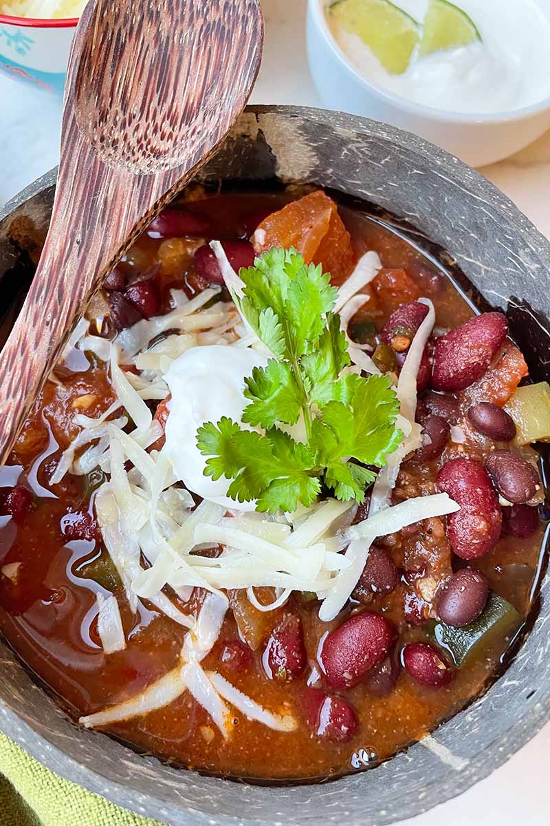 Vertical close-up image of the top of a wooden bowl filled with a chunky bean soup with a shredded white cheese and cilantro garnish, with a wooden spoon resting on the side.
