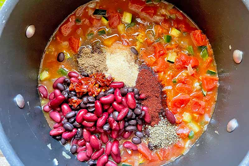 Horizontal image of assorted beans and spices on top of a tomato mixture in a pot.