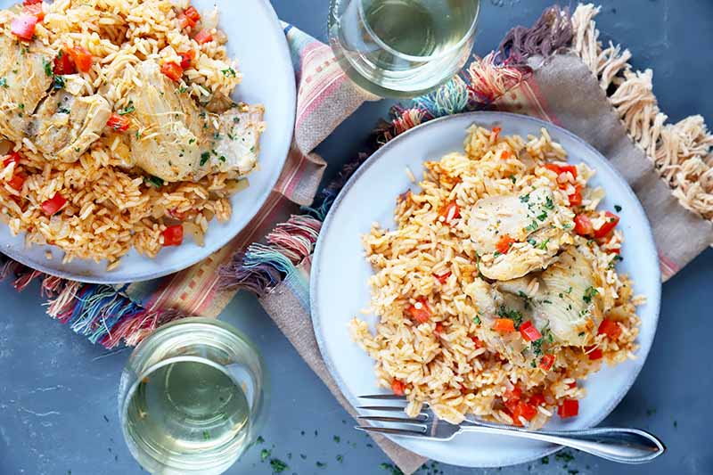 Horizontal image of two plates of chicken and rice next to glasses of white wine, a towel, and silverware.