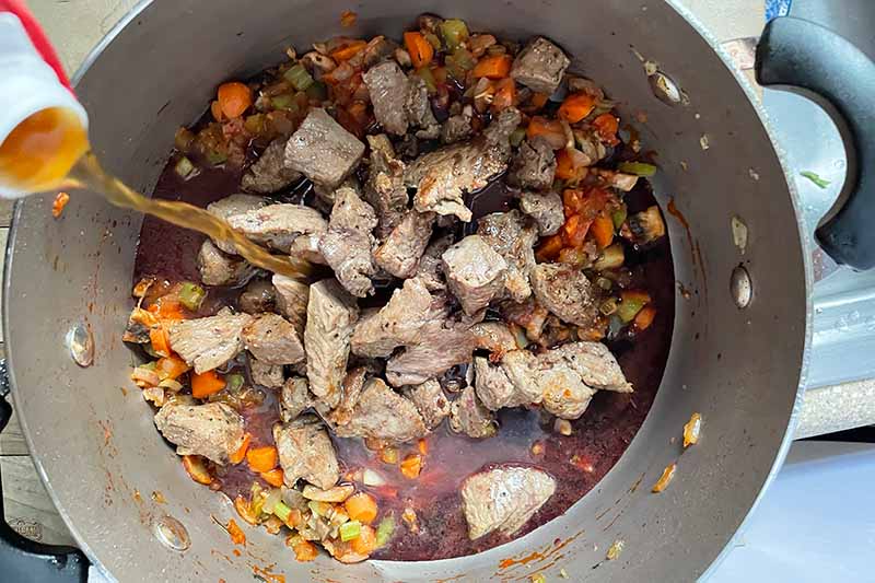 Horizontal image of adding stock to a pot with chuck roast chunks and chopped vegetables.