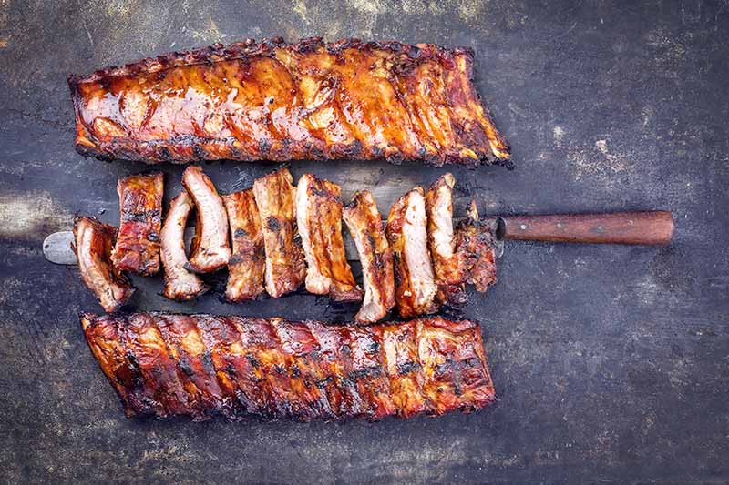 Horizontal image of three racks of bone-in barbecued meat, with one on a spike.