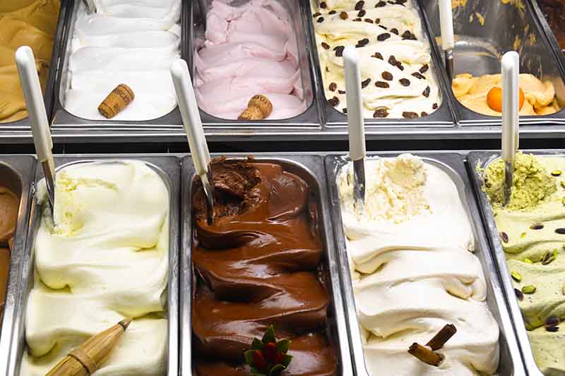 Horizontal image of trays of assorted gelato with scoops.