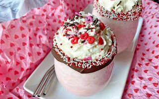 Horizontal image of two glasses with a creamy pink beverage garnished with whipped cream, chocolate rim, and sprinkles on a white plate.