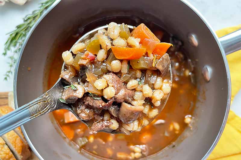 Horizontal image of a ladleful of a grain, meat, and vegetable stew over a metal pot.
