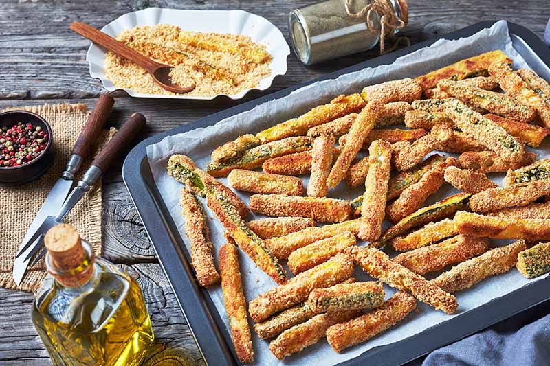 Horizontal image of seasoned and dredged zucchini sticks on a baking sheet next to ingredients in bowls and bottles.