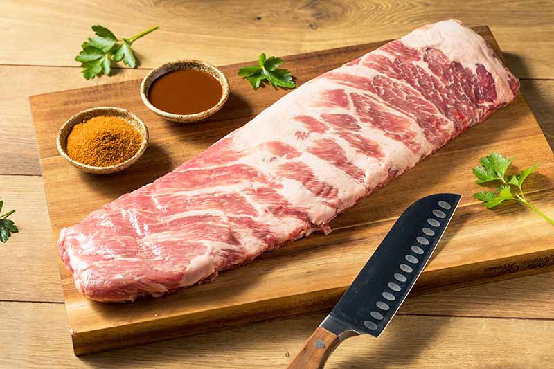 Horizontal image of a rare rack of pork meat on a wooden cutting board next to bowls of spices, fresh herbs, and a knife.