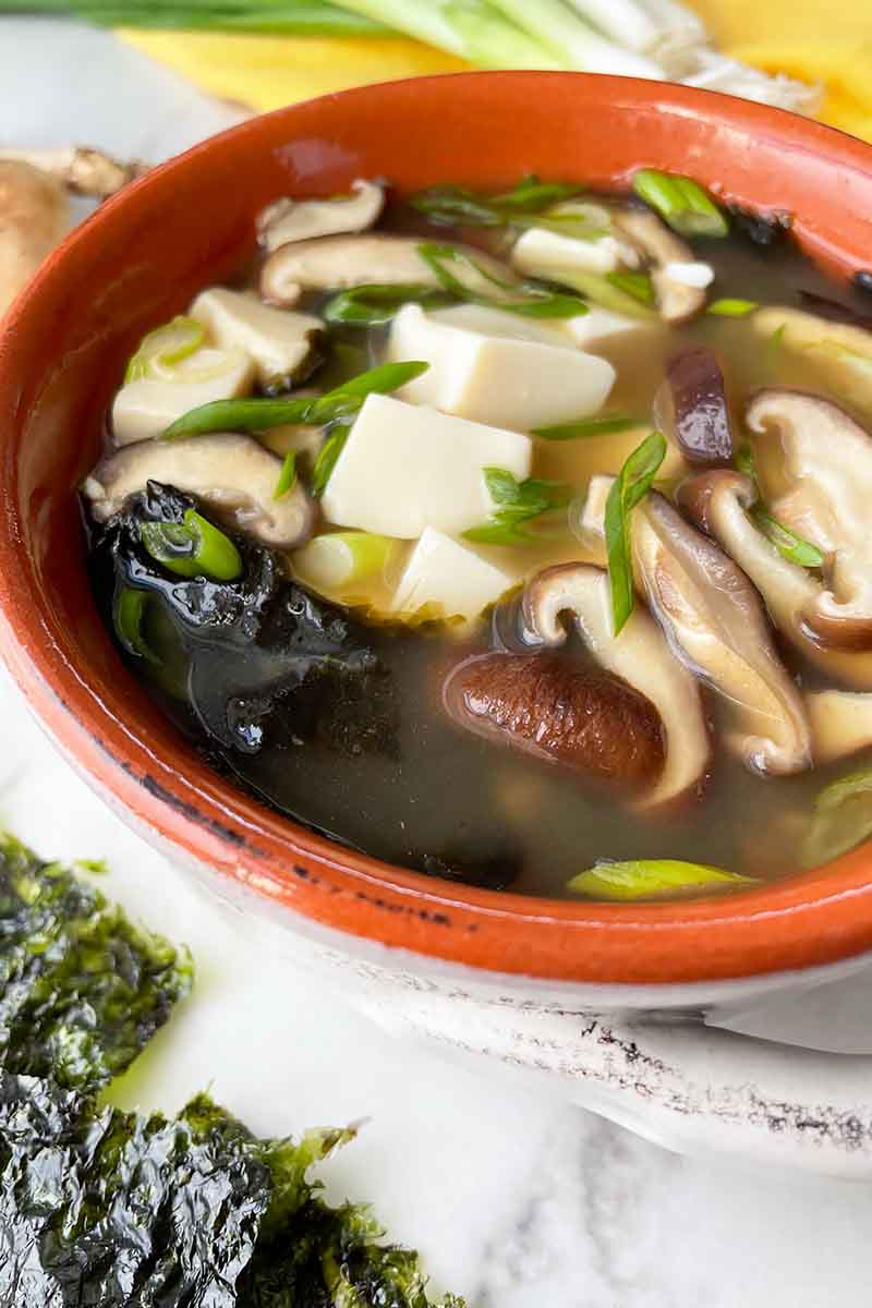 Vertical close-up image of a bowl full of broth with tofu chunks, sliced shiitake, and seaweed pieces.