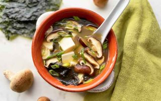 Horizontal image of a red bowl filled with broth with chunks of tofu, sliced mushrooms, and seaweed with a spoon inserted into it, next to a green towel