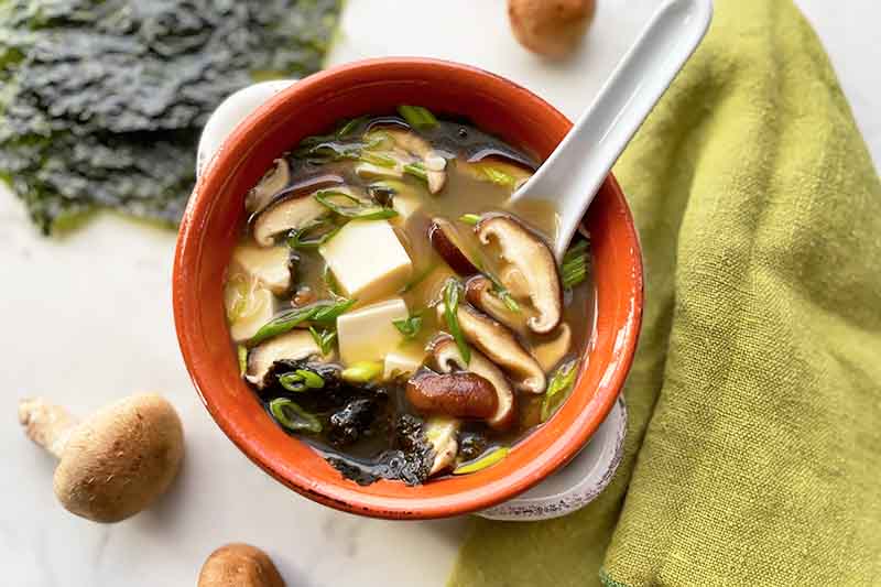 Horizontal image of a red bowl filled with broth with chunks of tofu, sliced mushrooms, and seaweed with a spoon inserted into it, next to a green towel