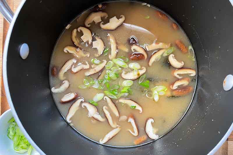 Horizontal image of a broth with sliced mushrooms and scallions in a black pot.