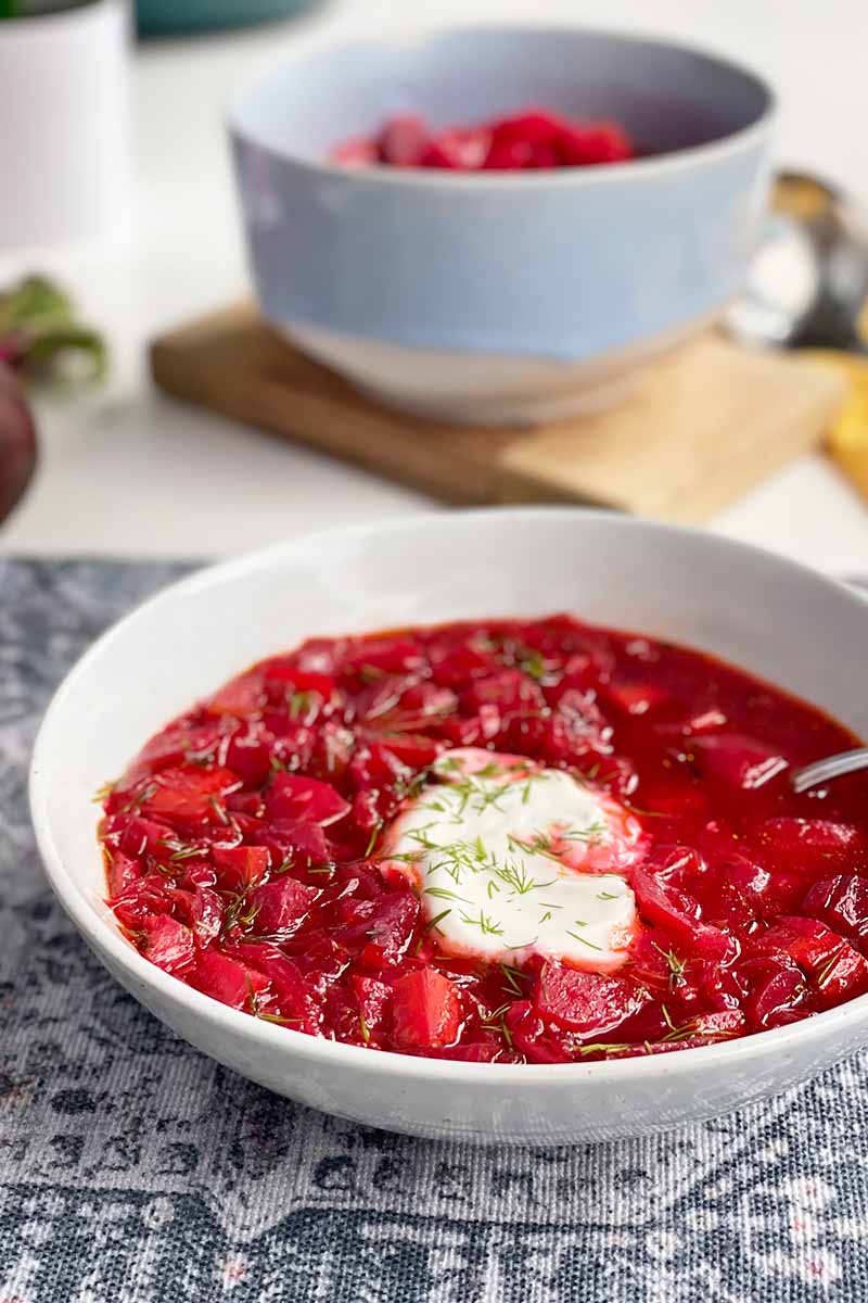 Vertical image of two bowls filled with borscht, with one garnished with sour cream and herbs on a navy towel.