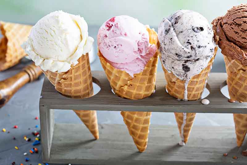 Horizontal image of scoops of assorted frozen desserts in a waffle cone in a stand.